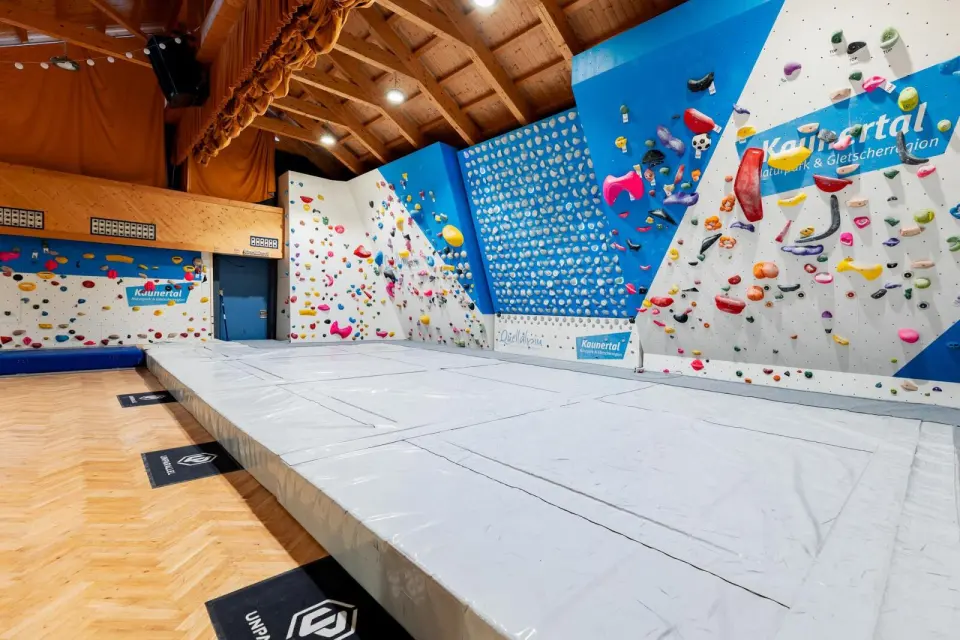 Blue and white bouldering walls and a Kilterboard above thick grey mats under a wooden roof