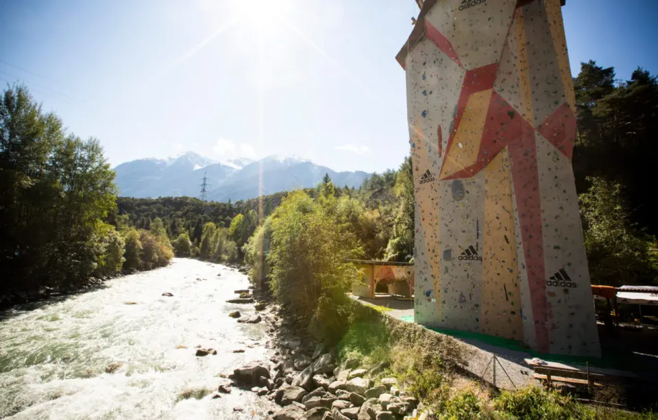 Tall outdoor climbing wall on a bridge pillar beside a rushing river, with mountains behind