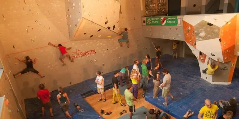 Climbers on the hall's tall walls, with an orange bouldering block and people on the mats