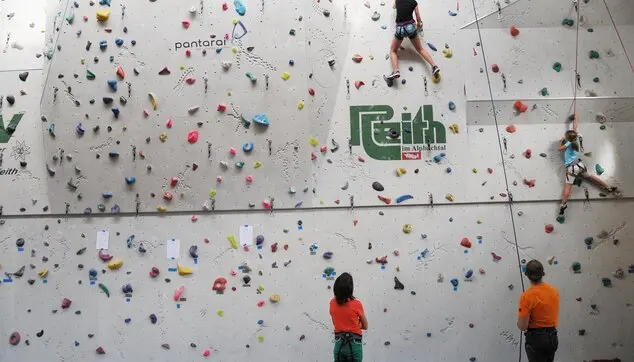 Climbers on top ropes high on a grey sports-hall climbing wall, with belayers below