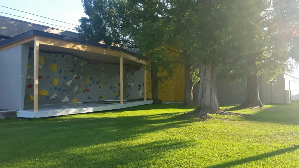 Bouldering wall under a timber roof on a sunny lawn, with a yellow lead wall behind the trees