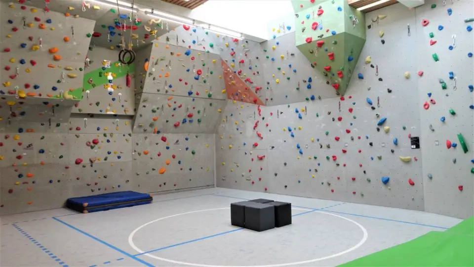 Grey lead walls with colourful holds, a green volume and quickdraws around an empty sports-hall floor