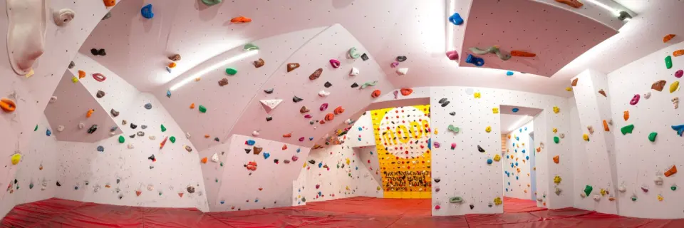 Bouldering room with white walls, colourful holds and red matting, a yellow training board at the back