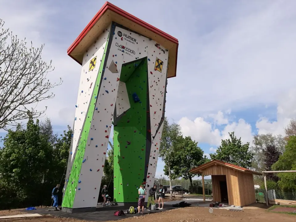The two-legged outdoor climbing tower with climbers at its base and a wooden hut beside it