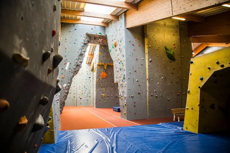Tall grey roped walls beyond a yellow bouldering wall and blue crash mats