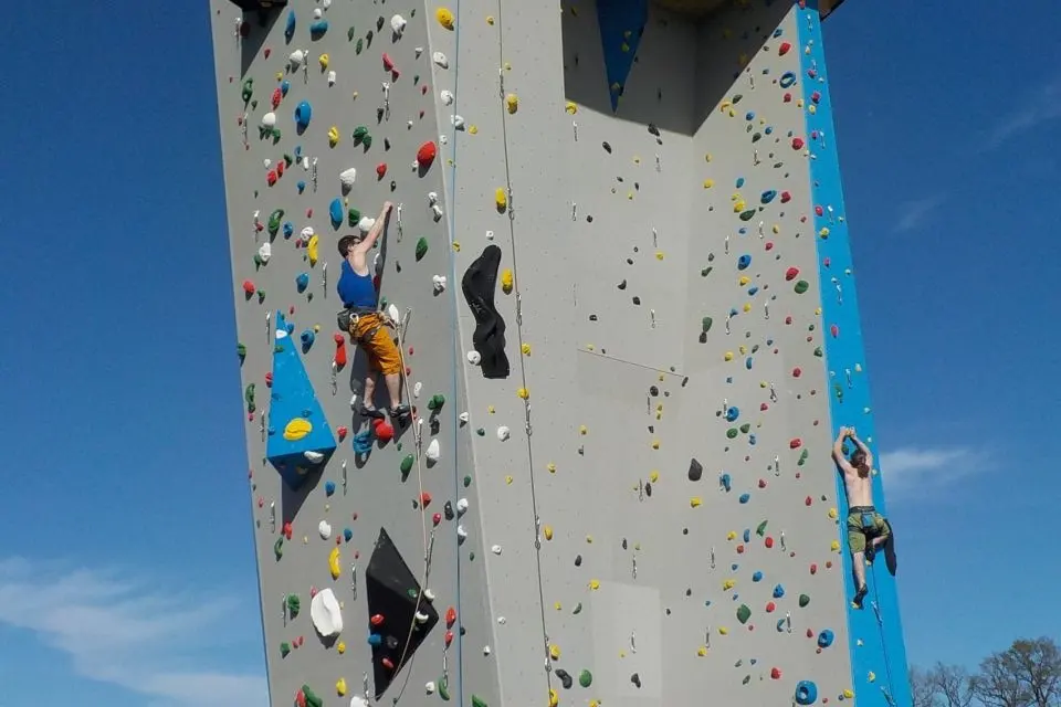 Climbers on the grey and blue outdoor climbing tower under a wooden roof