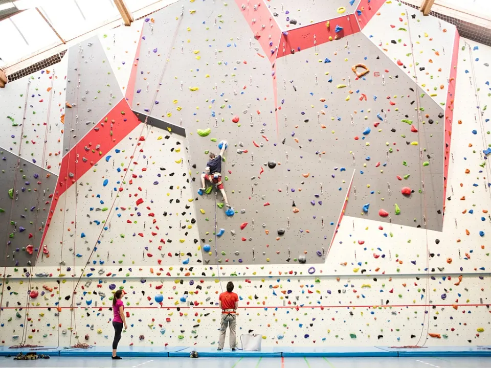 Climber high on a wide roped wall with grey and red panels while two people watch from the mats
