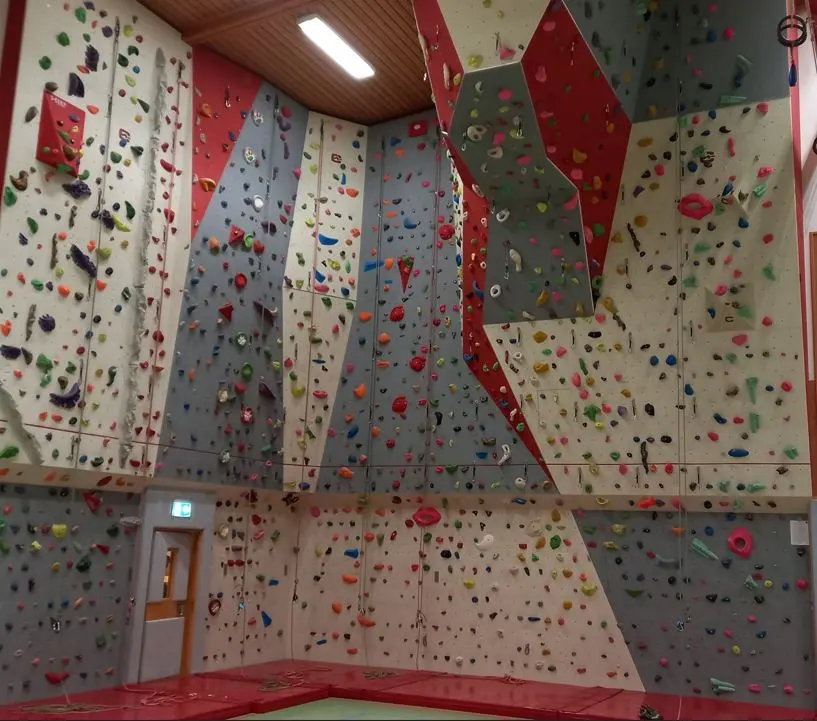 Corner of a roped climbing wall with grey, white and red panels, a roof section and red mats
