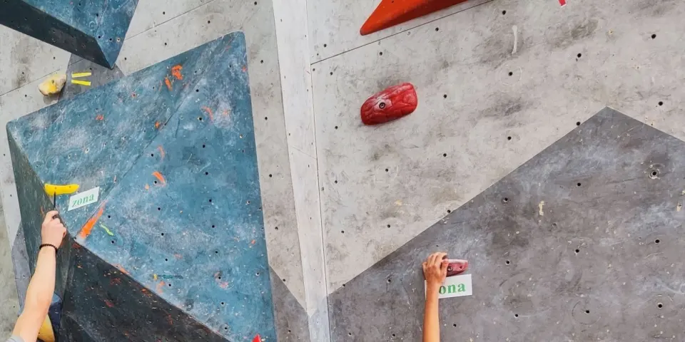 Climbers' hands on holds beside large blue volumes during a bouldering competition