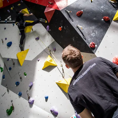Belayer watching a lead climber on a wall with yellow volumes and quickdraws