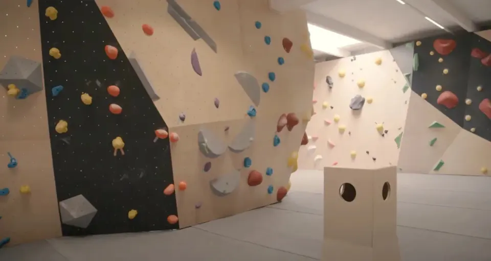 Plywood and black bouldering walls with coloured holds and grey volumes above thick matting