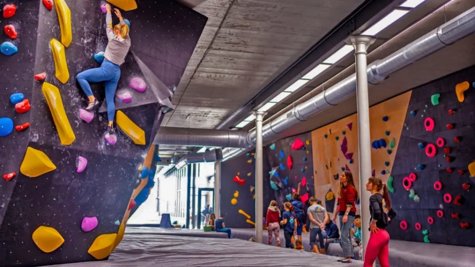 Climber on a steep wall while others gather on the mats in the long bouldering hall