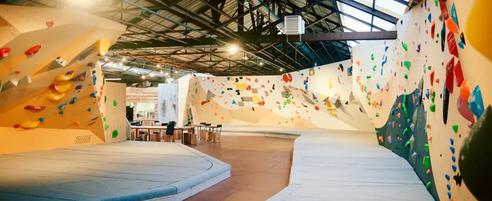 Panorama of the bouldering hall, with padded matting below the walls and the bar tables beyond