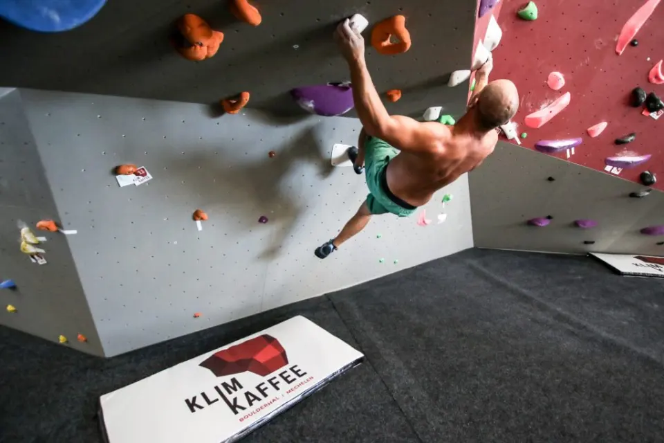 Climber on a grey overhang above a crash pad printed with the Klimkaffee Mechelen logo