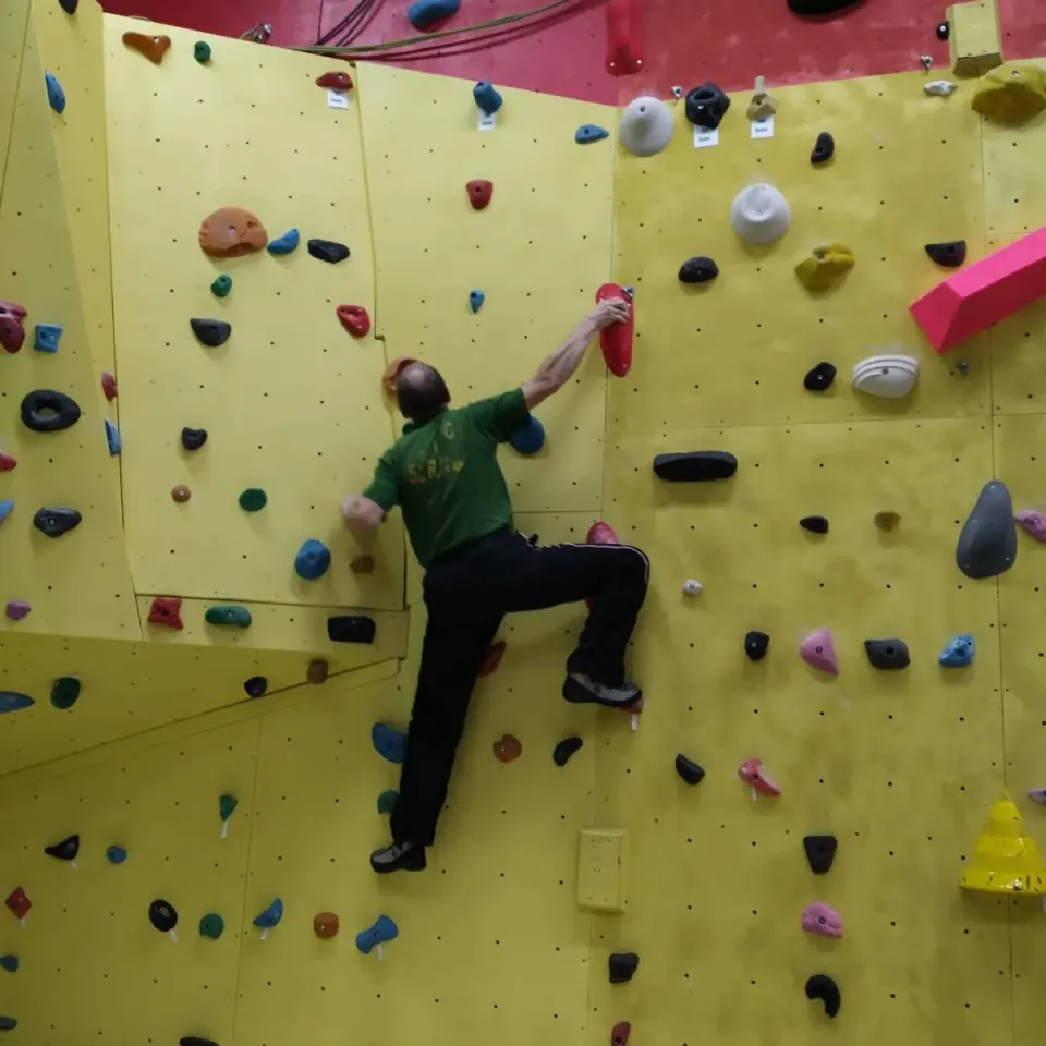 Climber in a club shirt reaching across the yellow bouldering wall