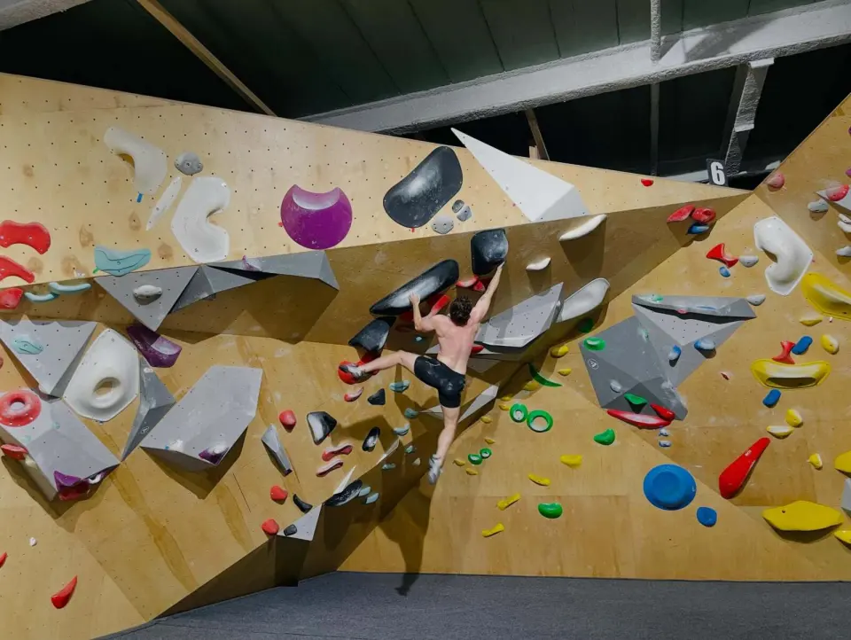 Climber on a plywood bouldering wall covered in grey volumes and colourful holds