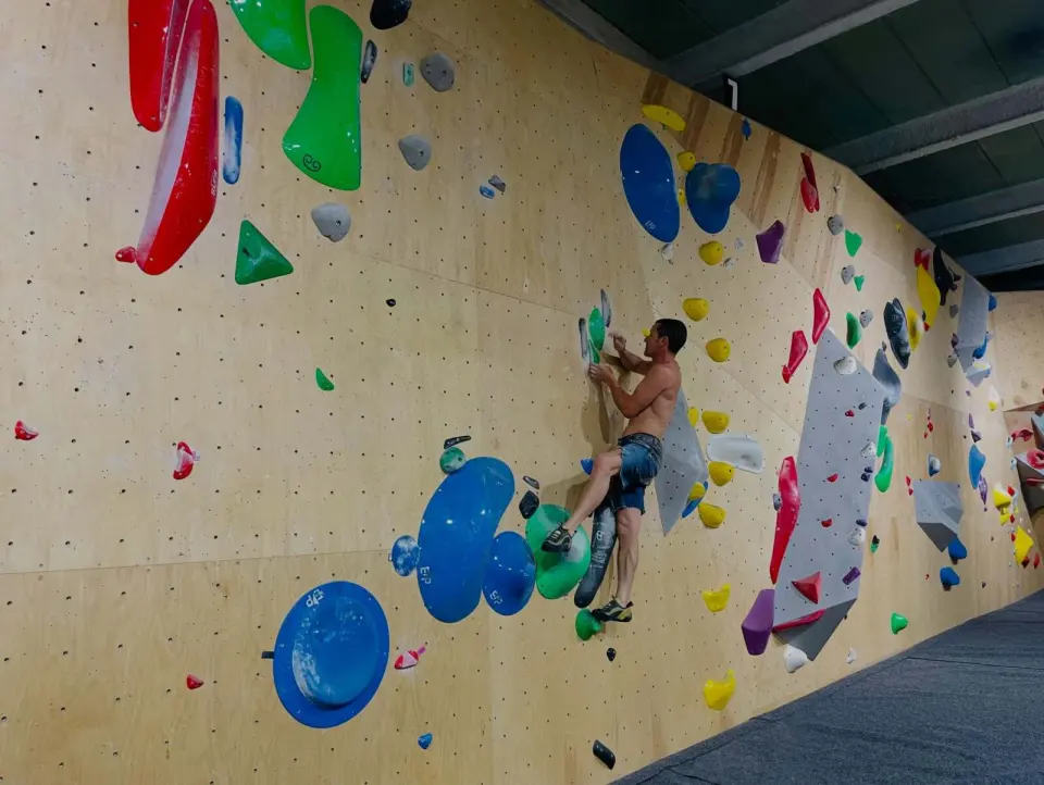 Climber on the long plywood wall, with big blue volumes and problems running along it