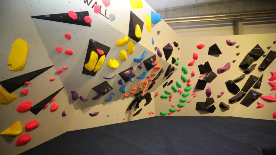 Long bouldering wall with angled panels, black volumes and bright holds above blue matting