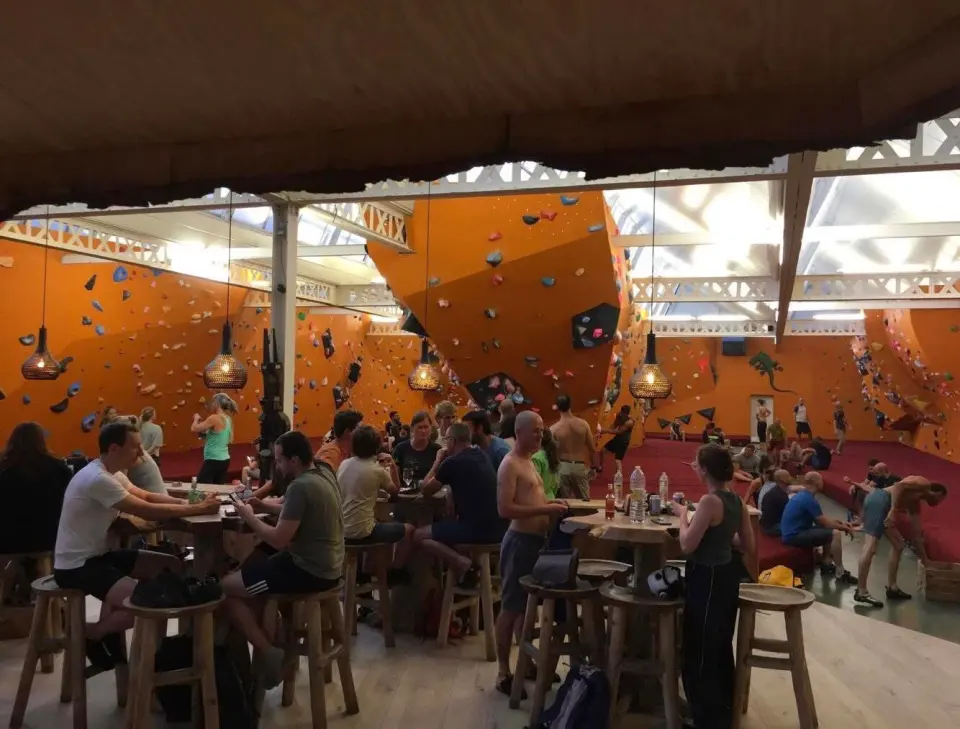 Busy bar tables in front of the orange bouldering walls and red mats under a glass roof
