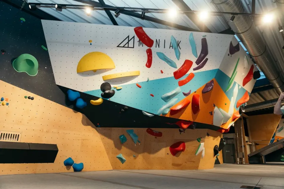 Overhanging bouldering wall with big coloured holds and the Maniak logo, under spotlights