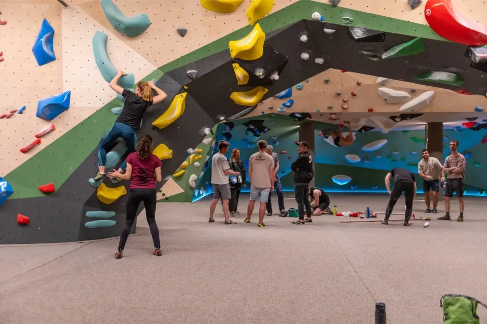 Climber on a green-edged overhang with big yellow holds while others gather on the mats