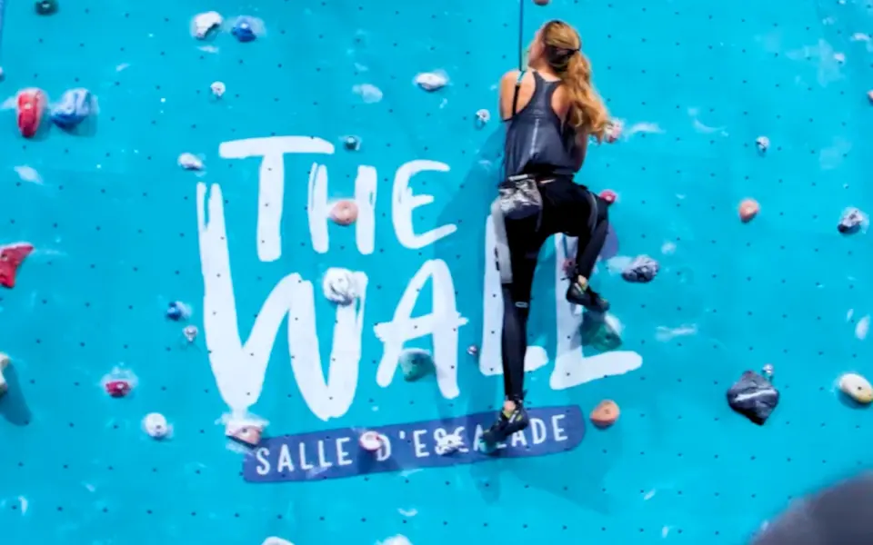 Climber on a top rope beside The Wall logo painted on the turquoise wall