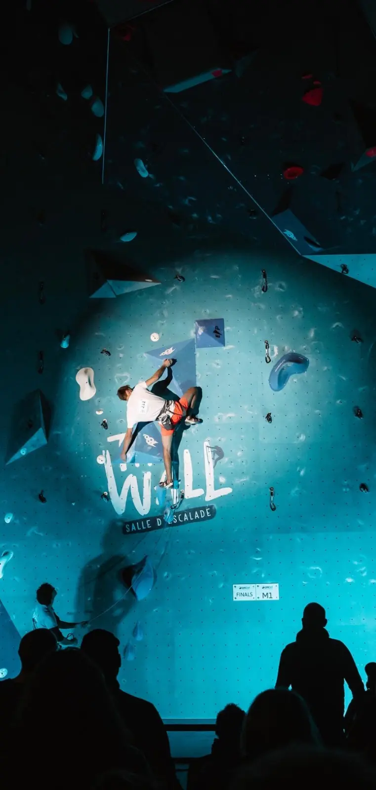 Lead climber in a competition final under a spotlight, watched by the crowd
