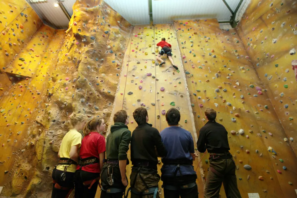 Climber on the yellow textured lead wall while a group watches from the matting
