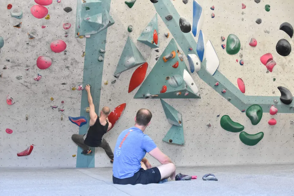 Climber on a wall of volumes while another climber watches from the mats