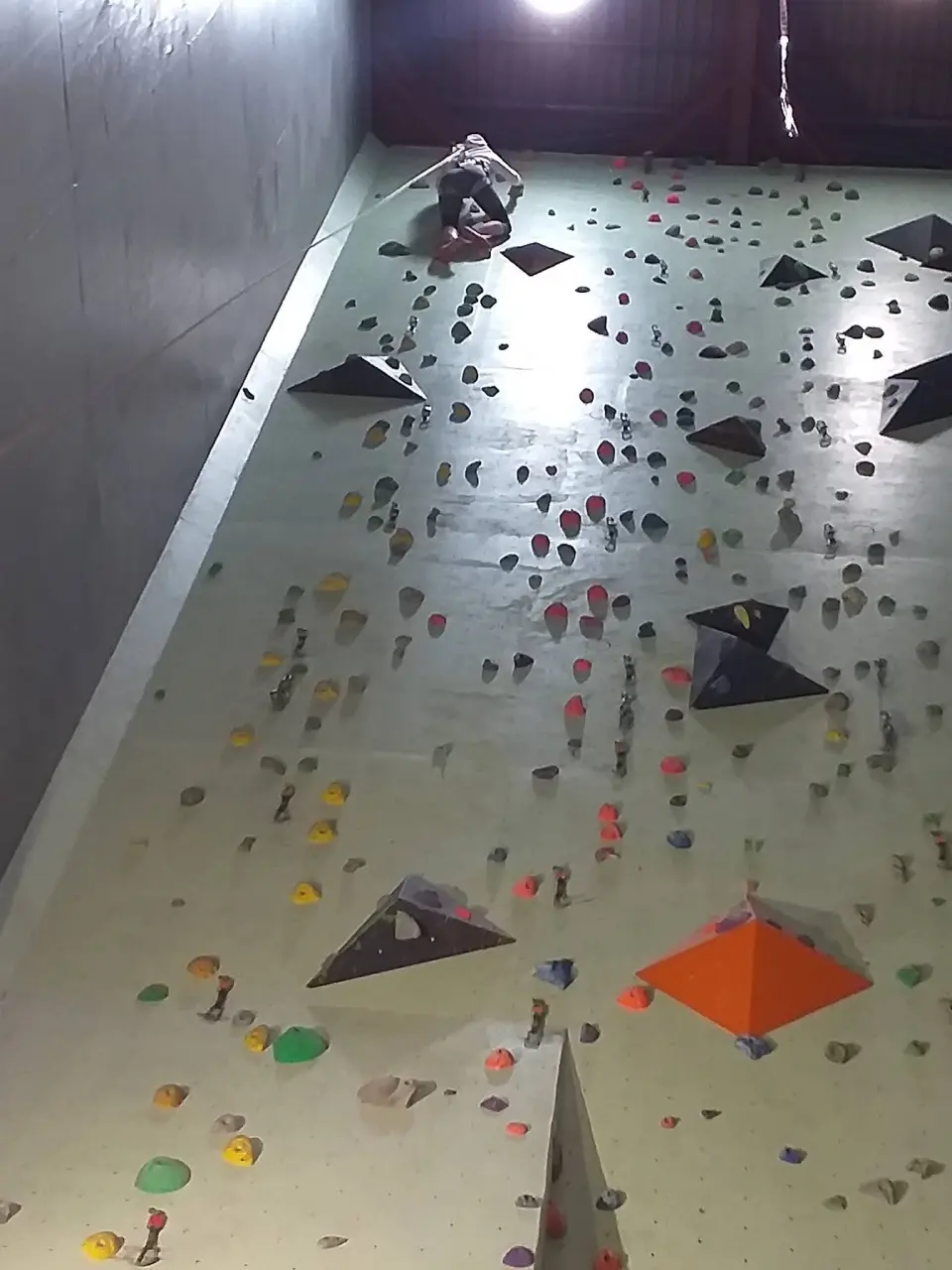 Tall indoor roped wall with volumes, looking up at a climber