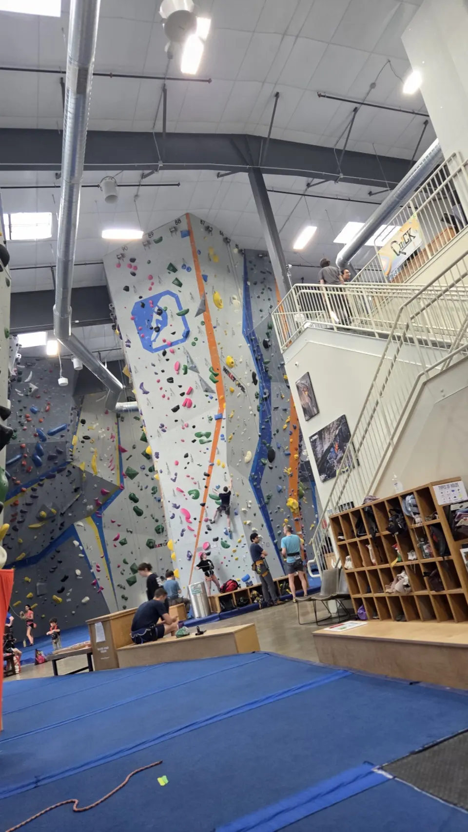 Climbers and belayers below the tall roped walls, with cubbies and the staircase