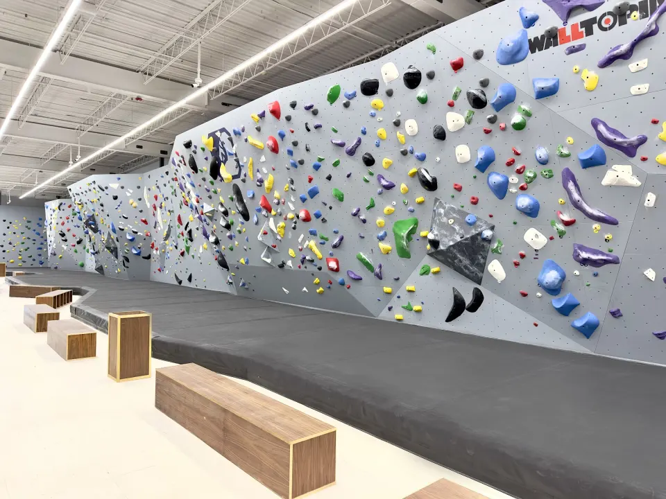 Long grey bouldering wall with colourful holds above thick black matting and wooden benches