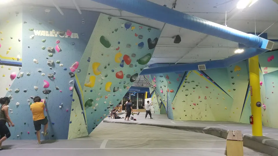 Teal bouldering walls under a blue air duct, with the yellow pillar and climbers in the background