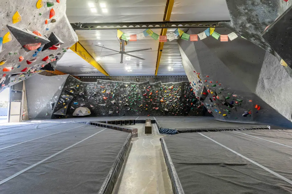 Bouldering hall with grey overhanging walls, yellow roof beams and grey matting