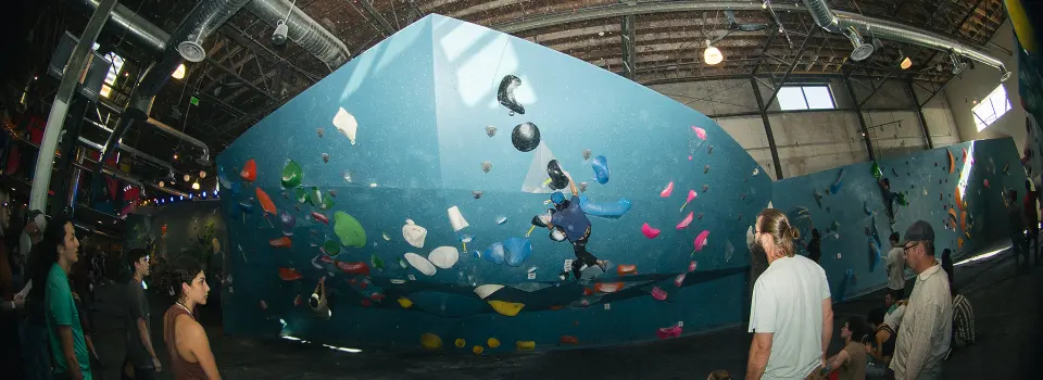 Wide view of a tall teal boulder with climbers watching from the mats