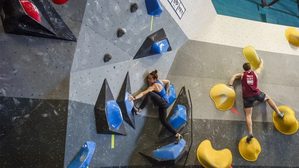 Two climbers on competition-style boulders with blue and yellow volumes
