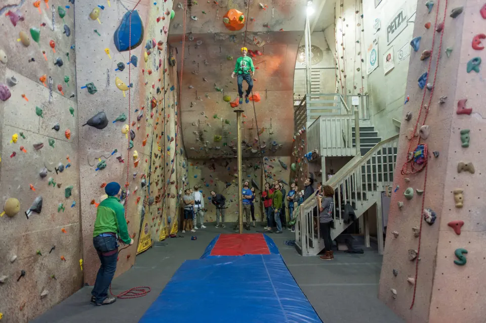 Tall roped wall above blue crash mats, with the stairway behind