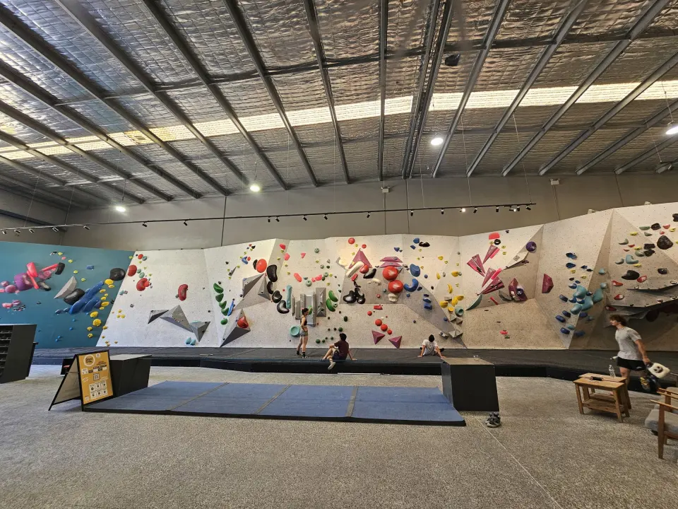 Long bouldering wall with teal and white panels under the warehouse roof