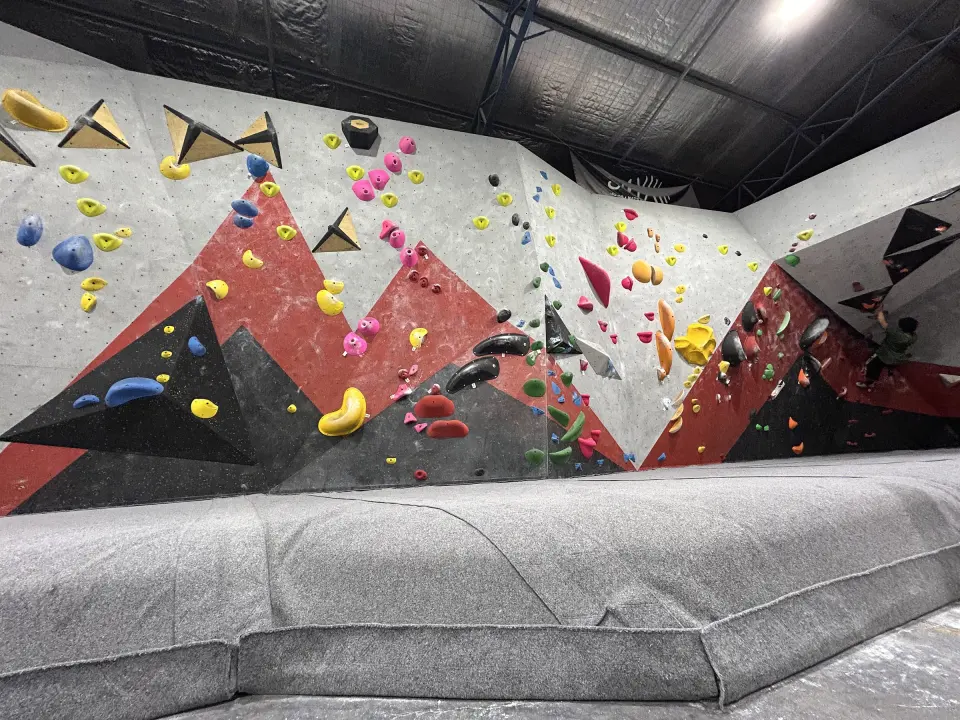 Long bouldering wall with angled red and black features and thick matting