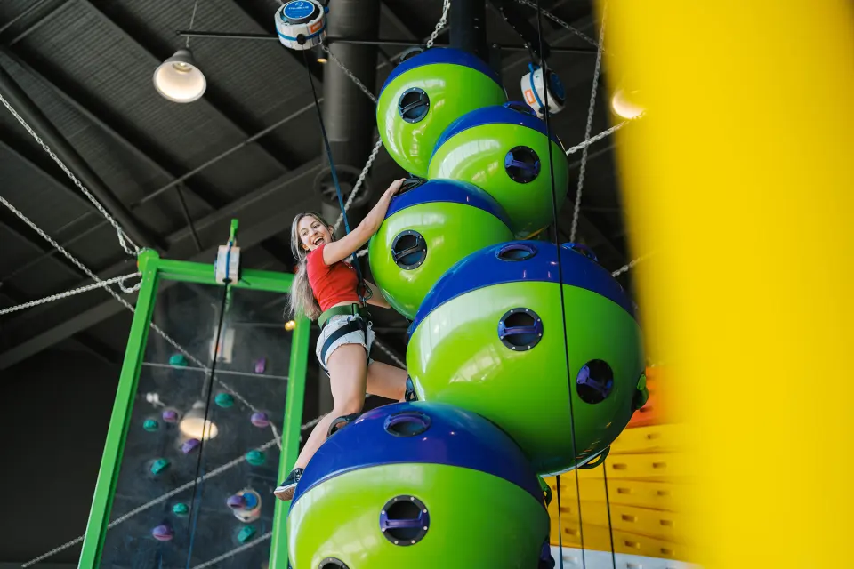 Climber on the stack of giant green and blue spheres beside a framed climbing wall
