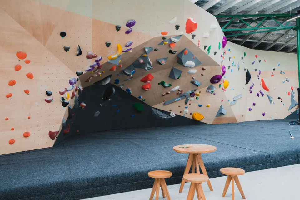 Overhanging bouldering wall above thick matting, with stools and a table in front