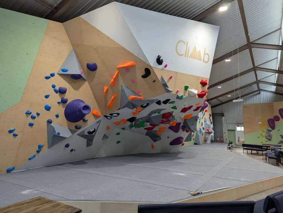 Bouldering wall with grey volumes and the Climb logo above padded matting