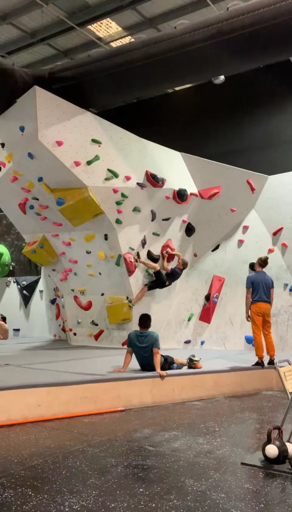 Main bouldering wall with yellow and red volumes and climbers resting on the mats