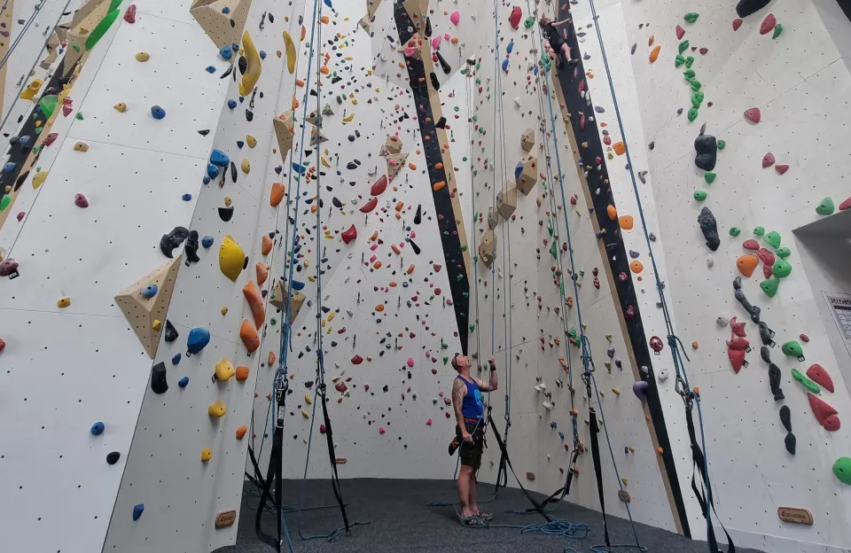 Belayer at the foot of the tall white rope walls, with quickdraws and ropes running up them