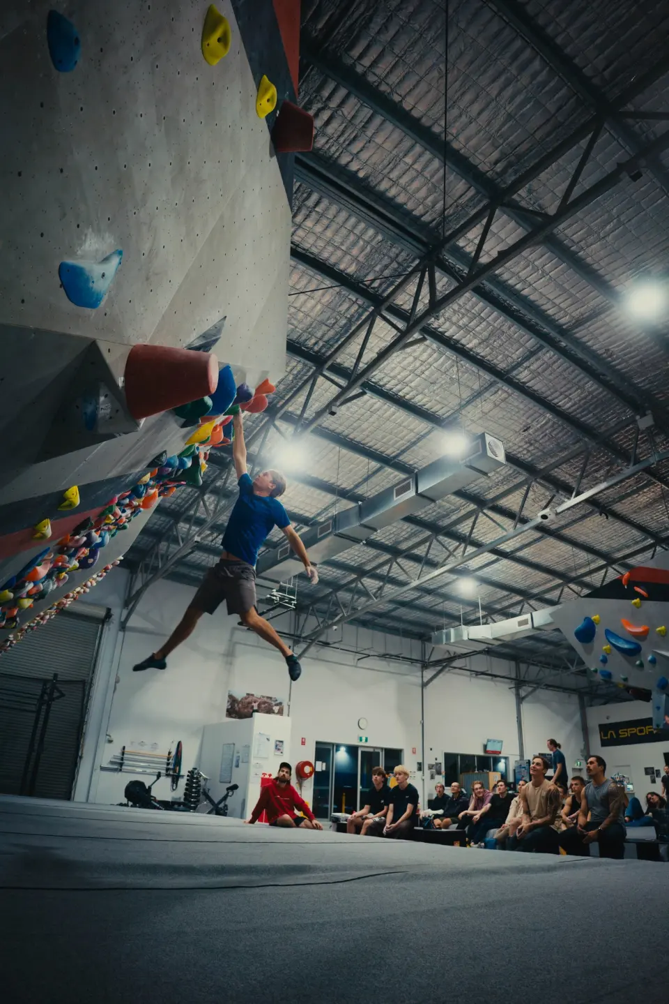 Climber jumping for a hold on the steep wall while a crowd watches from the mats