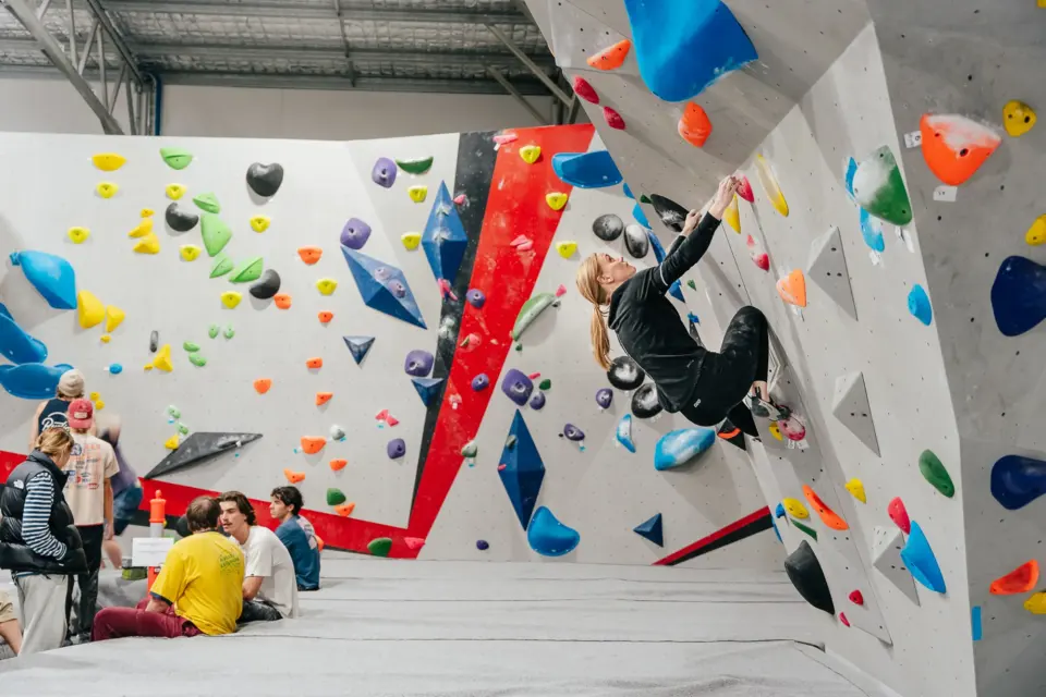 Climber on a vertical wall with a red stripe as others rest on the matting