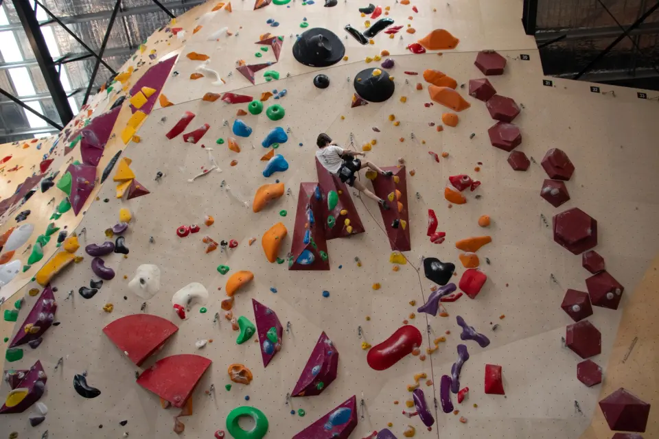 Climber on a tall rope wall covered in coloured holds and volumes