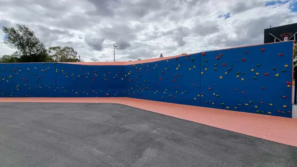Long blue outdoor climbing wall with coloured holds above orange rubber surfacing