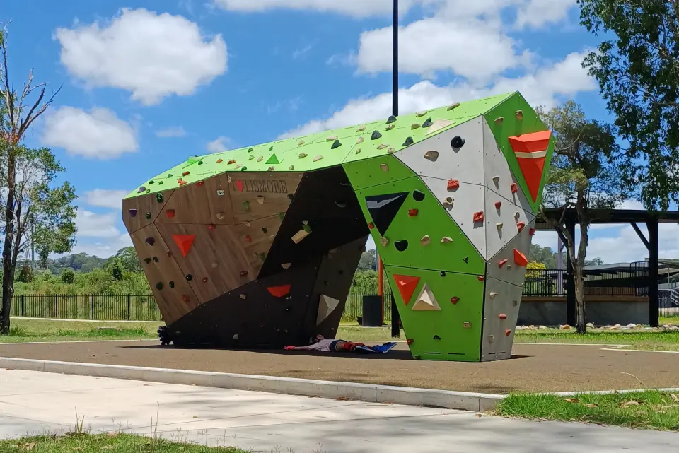 Green and timber-look bouldering block with an overhanging arch, on softfall beside a park path