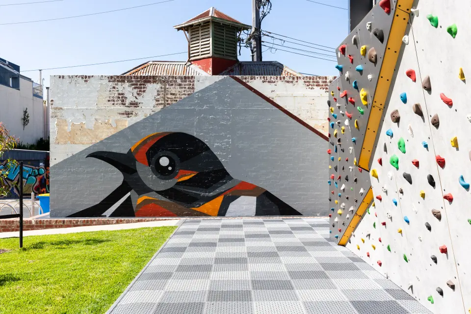 Bouldering wall covered in coloured holds above grey rubber tiles, with a bird mural on the brick wall beyond